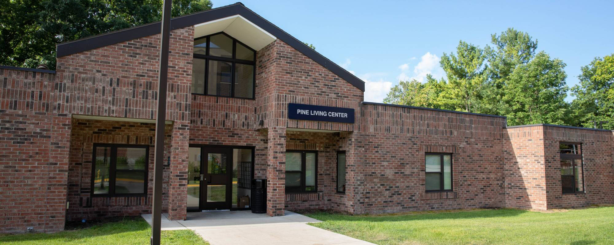 Red-brick building with dark trim labeled "Pine Living Center" against a clear blue sky. Surrounded by green lawn and trees, with a welcoming entrance.